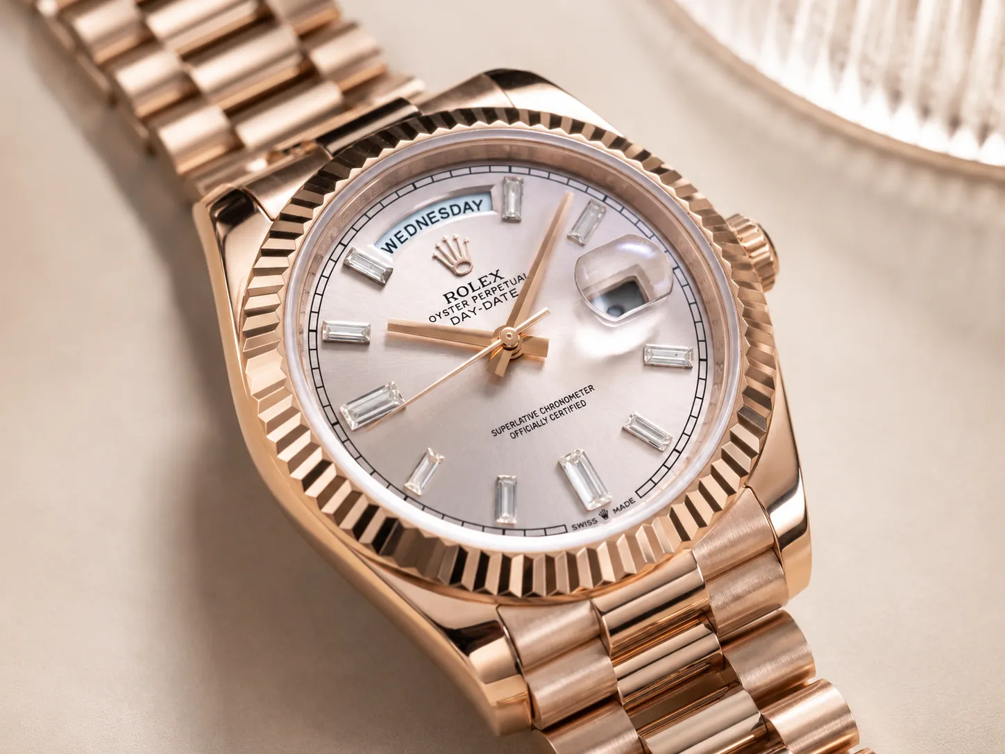 Close-up of a rose gold Rolex Day-Date watch featuring a silver sunburst dial, fluted bezel, baguette-cut hour markers, day display at 12 o'clock, and magnified date window in an elegant luxury setting.